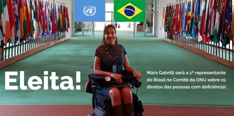 Descrição da imagem: A deputada federal Mara Gabrilli esta sorrindo numa cadeira de rodas ao centro de um salão com bandeiras nas lateriais, tendo ao fundo as imagens das bandeiras da ONU e do Brasil.