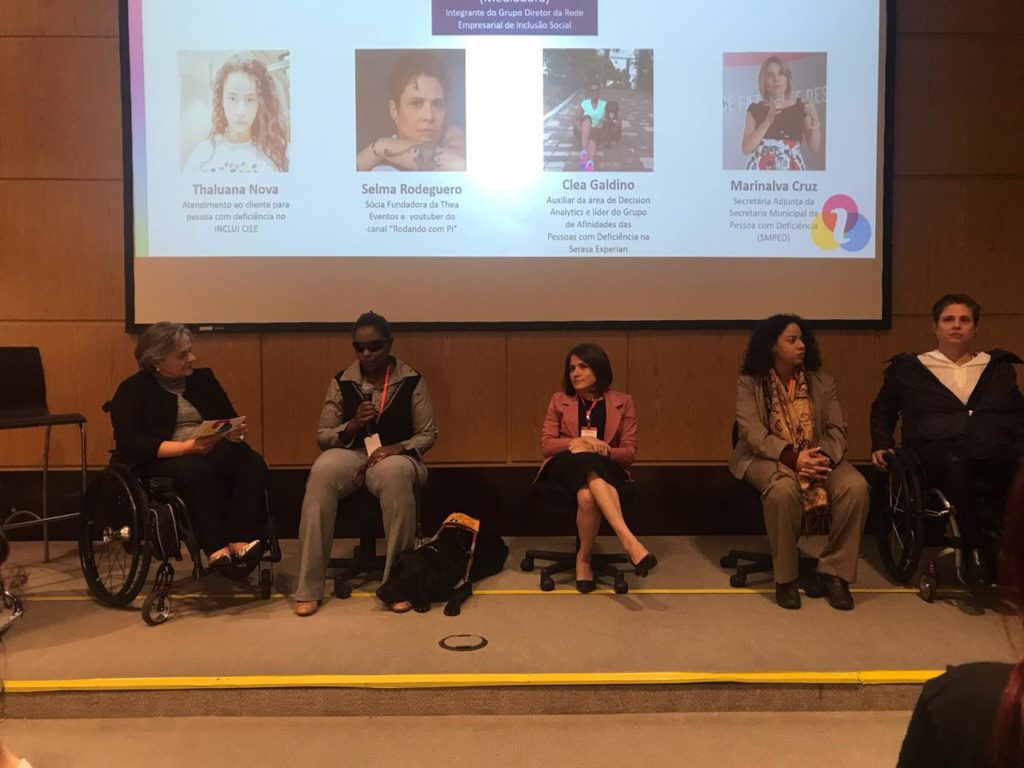 Descrição da Imagem: cinco mulheres sentadas no palco sob um painel com suas fotos. As participantes são Eliane Pelegrini, Clea Galdino, Marinalva Cruz, Thaluana Nova e Selma Rodeguero.