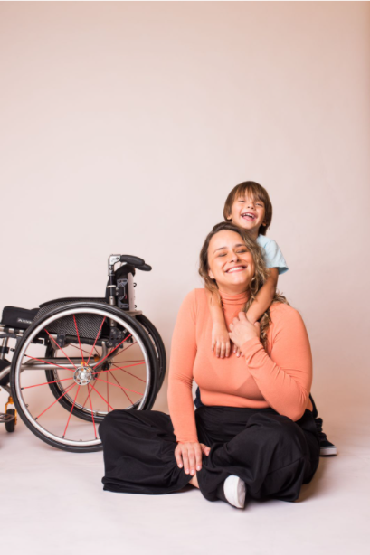 mulher branca, loira, sentada no chão, um menino pequeno a abraça por trás. ao lado esquerdo está uma cadeira de rodas.