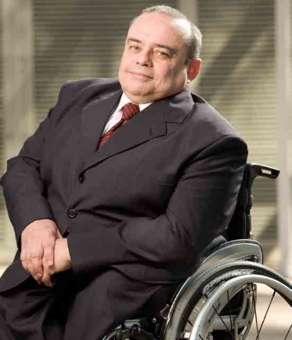 Homem branco, de cabelos curtos e castanhos e olhos castanhos. Ele está sentado em uma cadeira de rodas e veste um terno cinza com uma camisa branca por baixo e gravata vermelha.